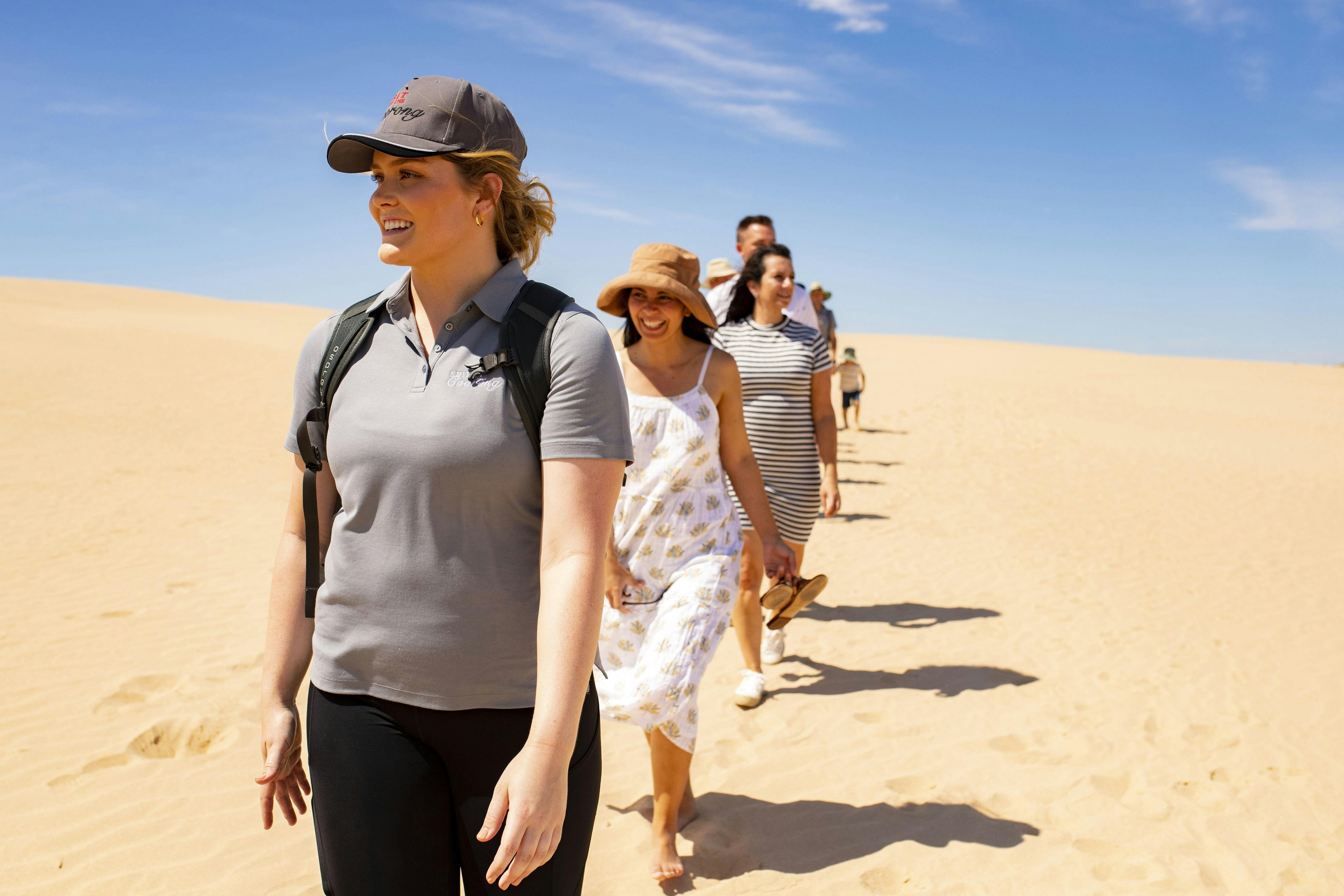 A group walking barefoot across sandy dunes under a bright blue sky, led by a smiling guide.