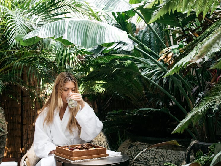 A girl sits in a white robe drinking a drink surrounded by plants and greenery