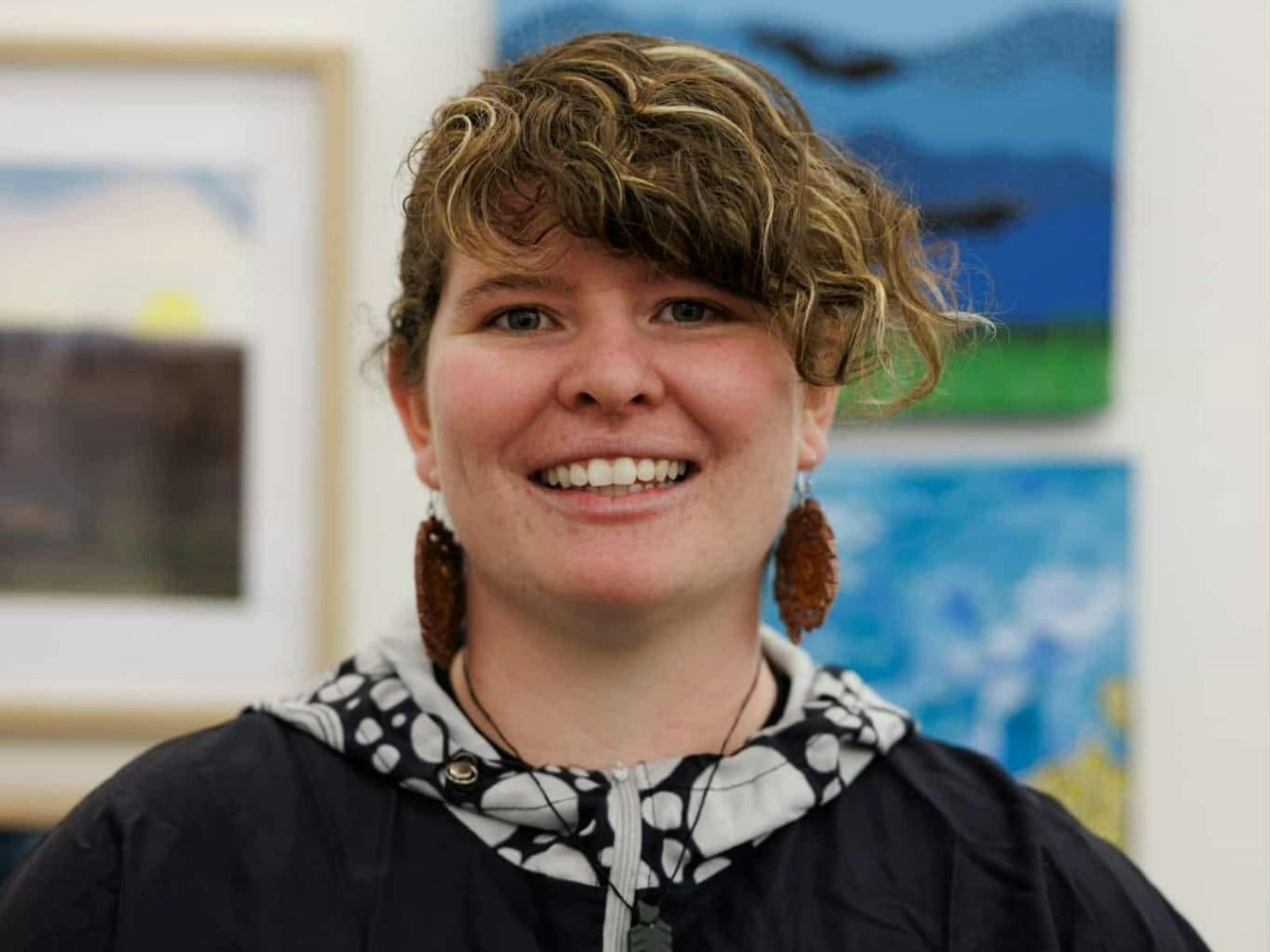 A smiling woman with curly brown hair and large brown earrings, wearing a black jacket.