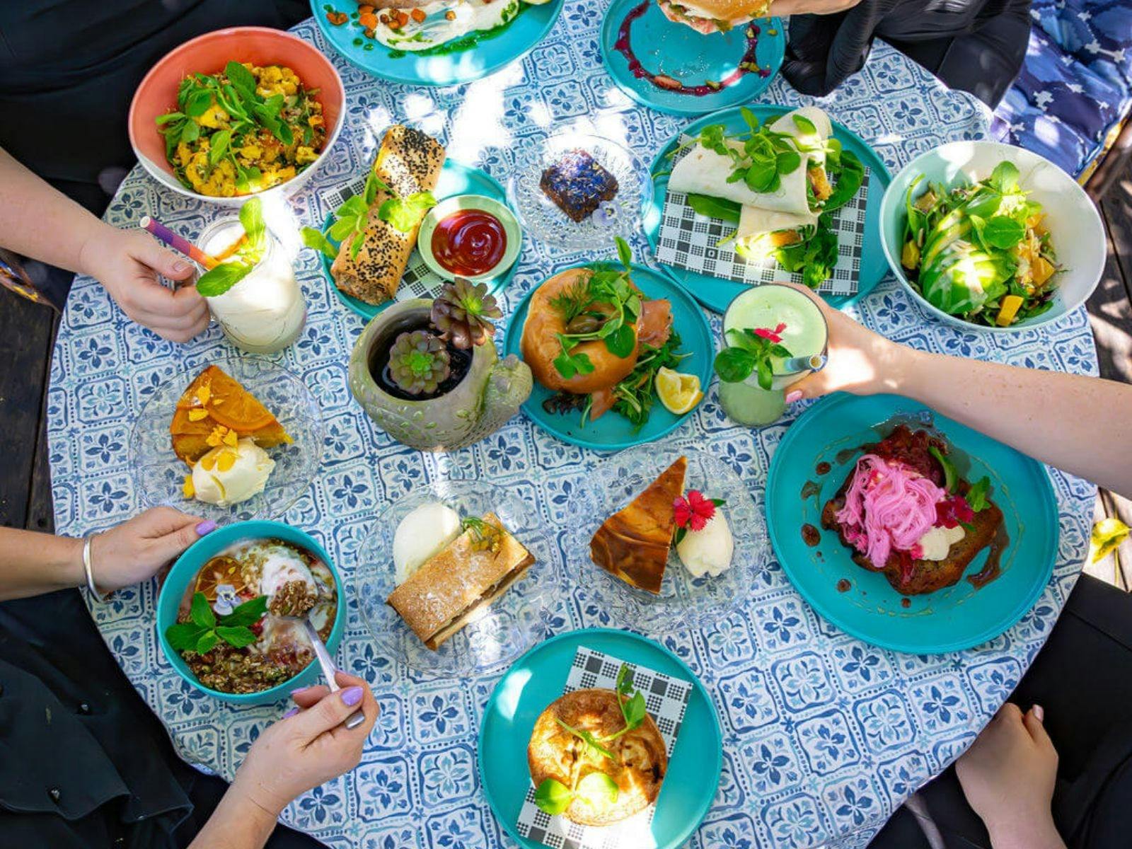 Overhead view of assorted colourful dishes and drinks on a patterned tablecloth, hands reaching in.