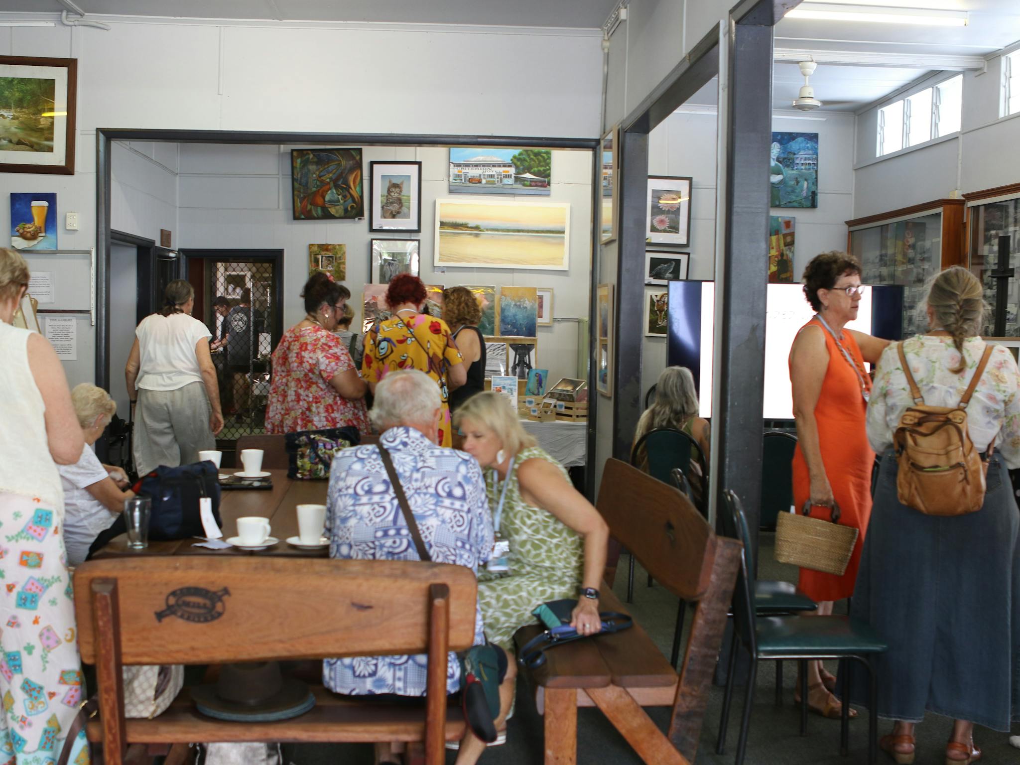 A group of people looking at artwork on the walls of a public venue. Some sits in front of tea cups. A group of people looking at artwork on the walls of a public venue. Some sits in front of tea cups.