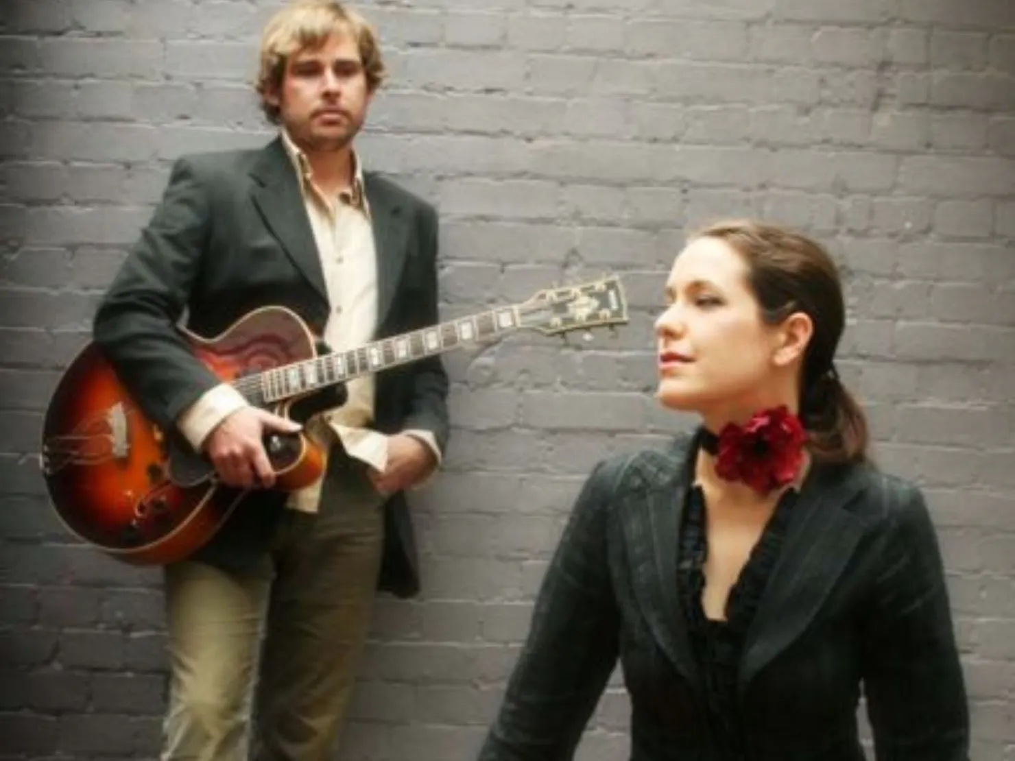 man with guitar in background with women in front with red rose around her neck