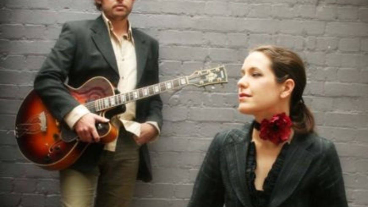 man with guitar in background with women in front with red rose around her neck