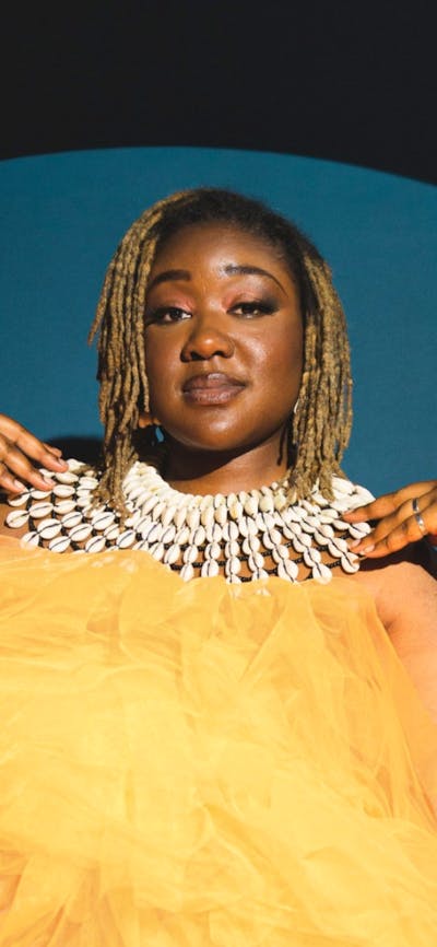 A woman sits in a yellow dress and white necklace wearing sneakers with