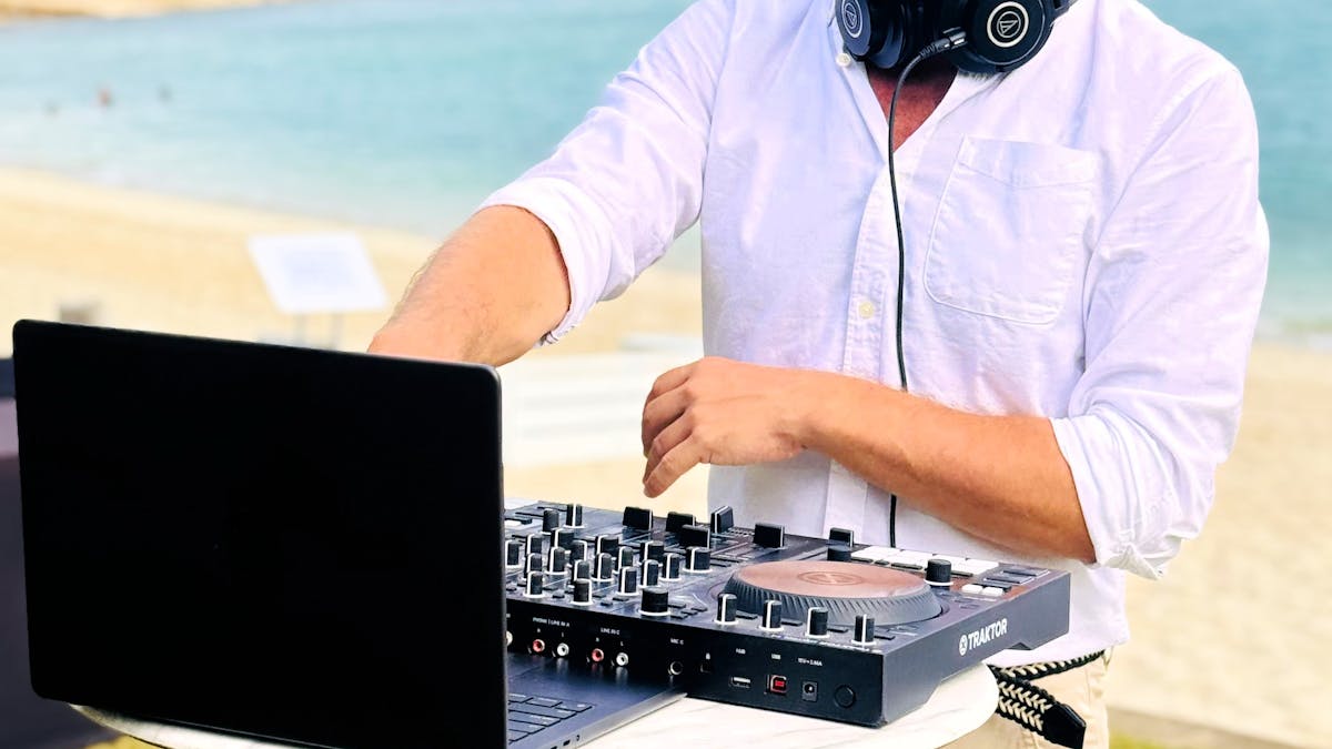 Image of a male DJ with their decks on a high bar performing with Hayman beach in the background