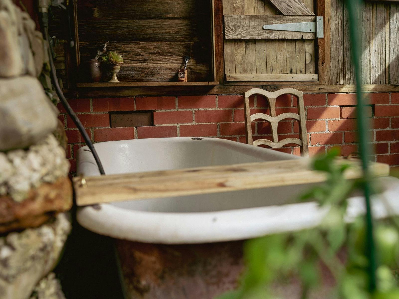 A rustic bathtub with vintage chair, brick wall, and timber window shutters.