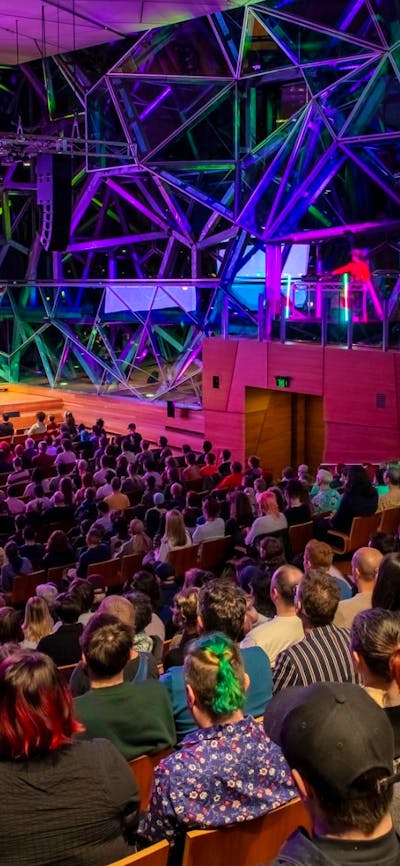 A large audience sits watching a stage with three people near a podium and a purple powerpoint slide