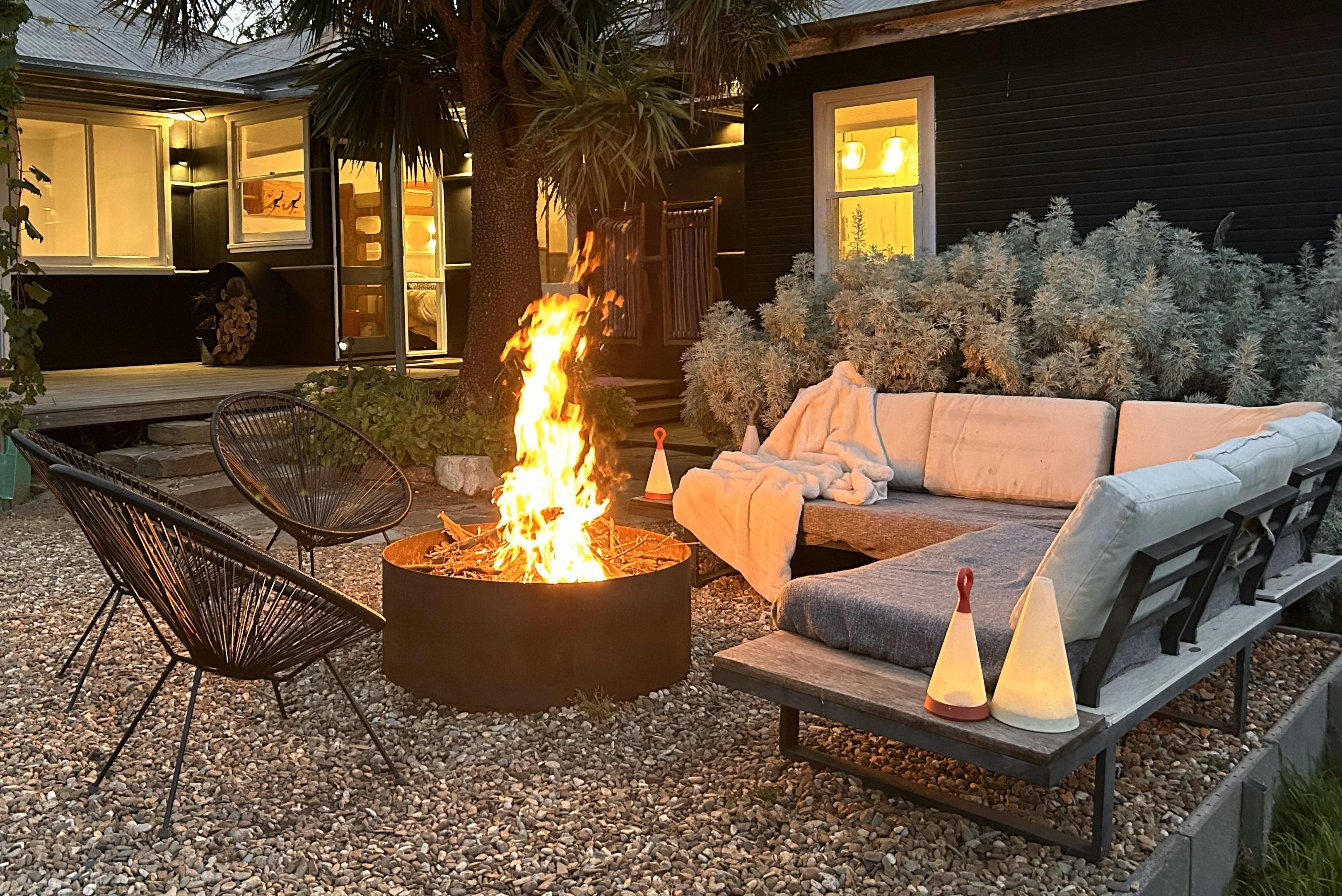 Firepit at dusk with outdoor couch, house in the background with lights on.