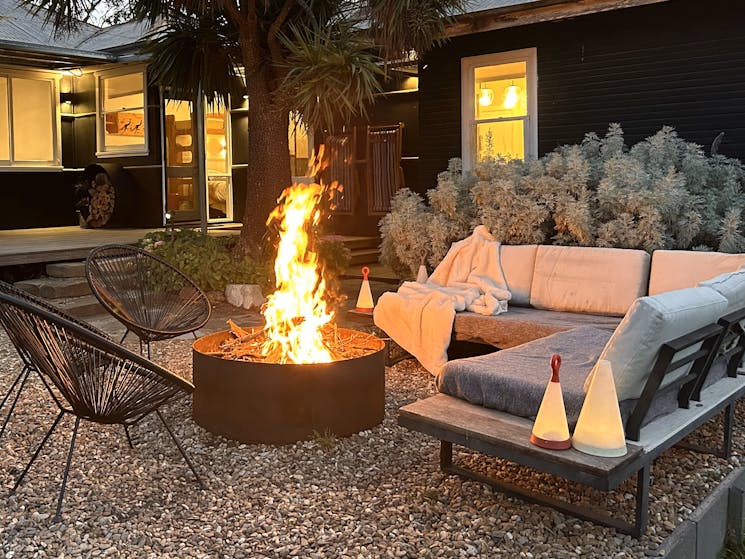 Firepit at dusk with outdoor couch, house in the background with lights on.