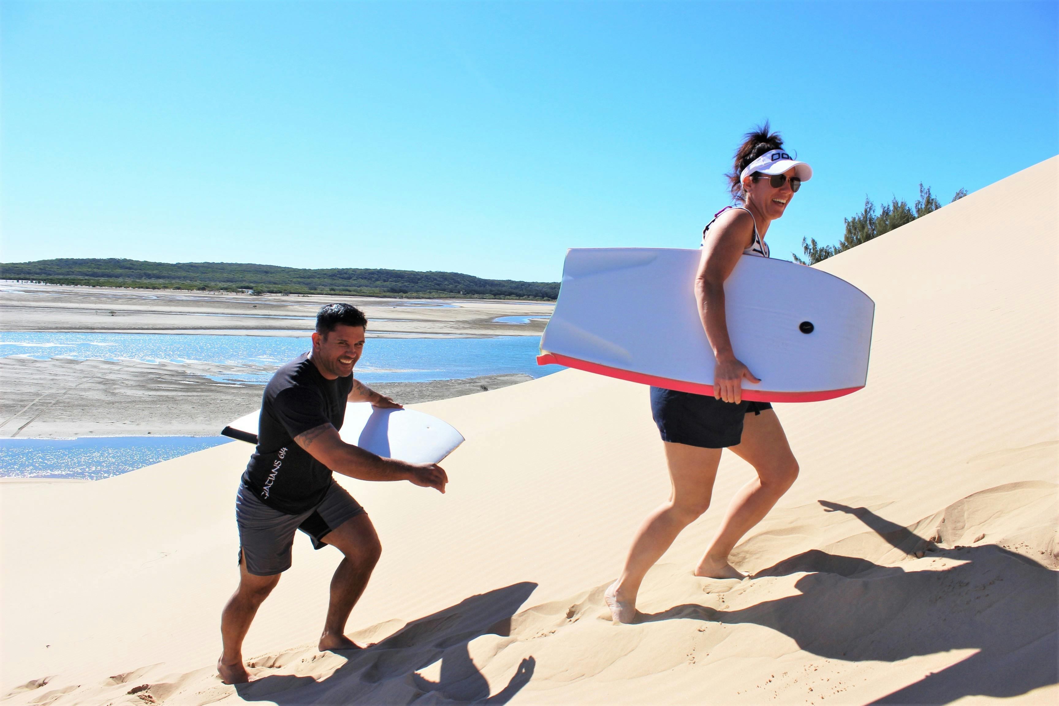 A man and a woman walking up a sand dune carrying body boards