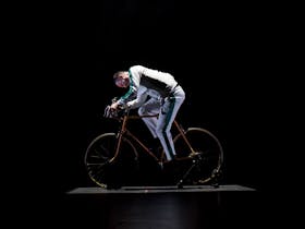 A man in a white tracksuit is riding a bicycle under a spotlight against a black backdrop