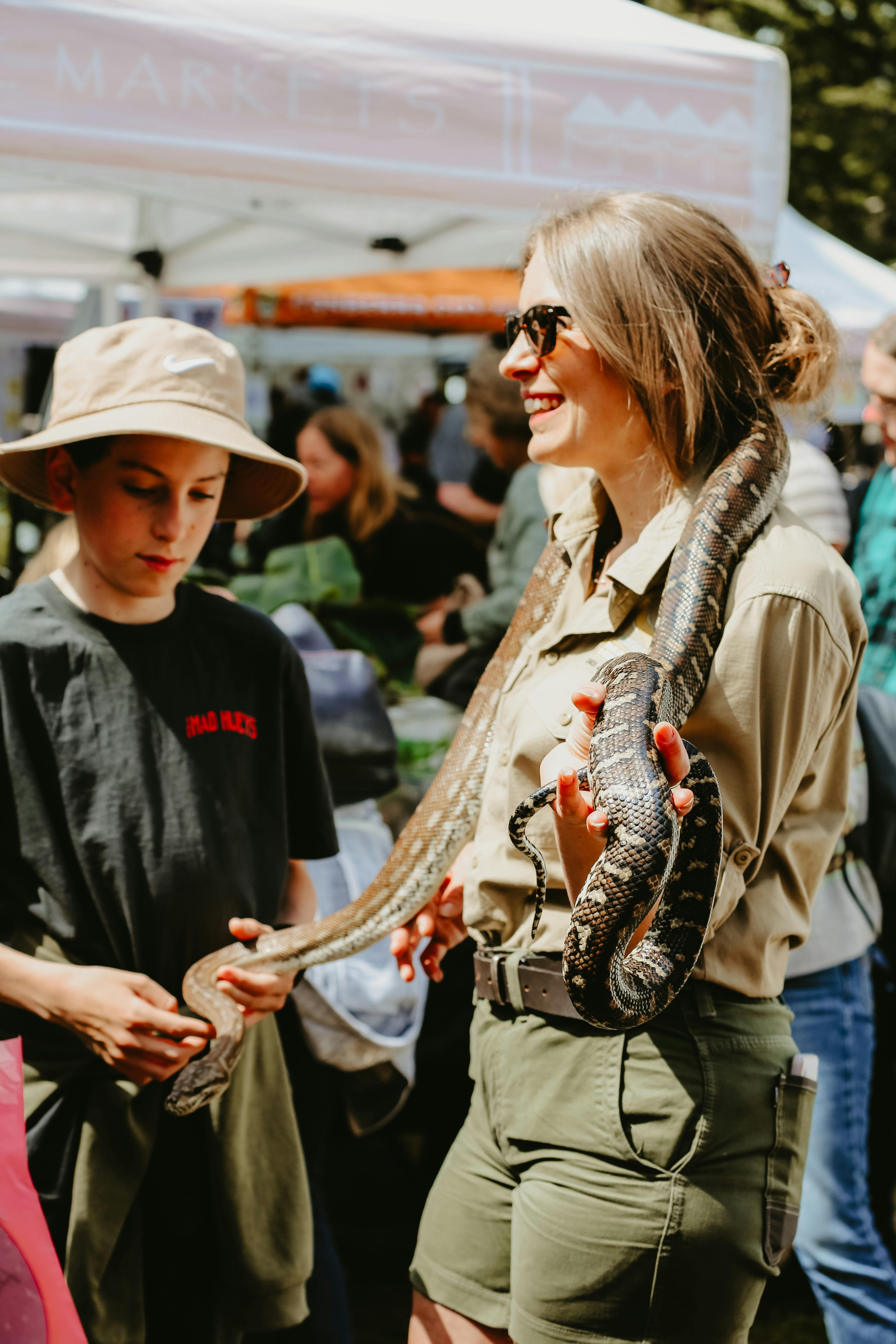 An image of a lady with a snake around her neck. A child nearby pats the snake.