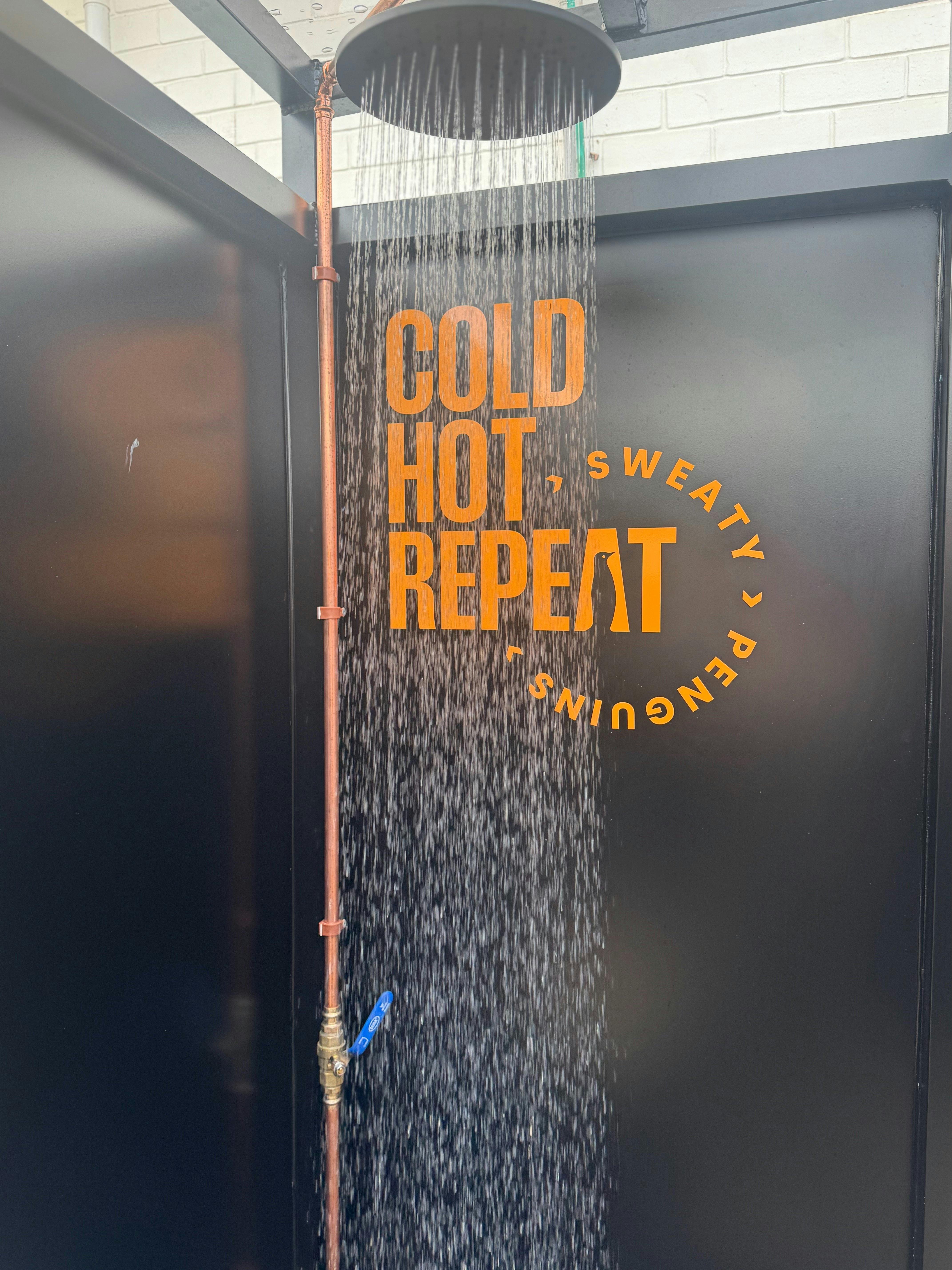 Cold outdoor shower with water running and 'Cold Hot Repeat' signage on the wall.