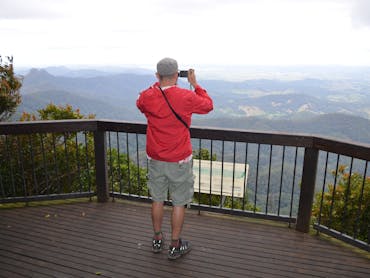 Man in red jacket looks thorugh binoculars at a lookout with views of mountains and valley.