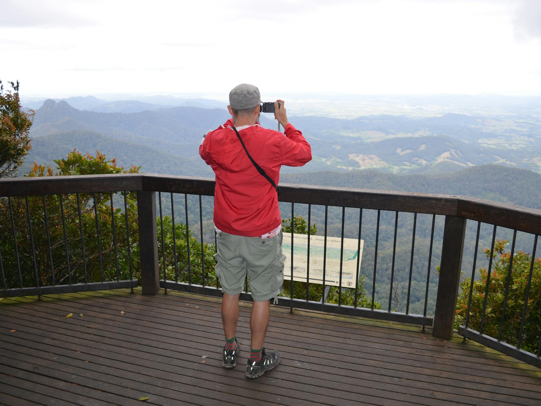 Man in red jacket looks thorugh binoculars at a lookout with views of mountains and valley. Man in red jacket looks thorugh binoculars at a lookout with views of mountains and valley.
