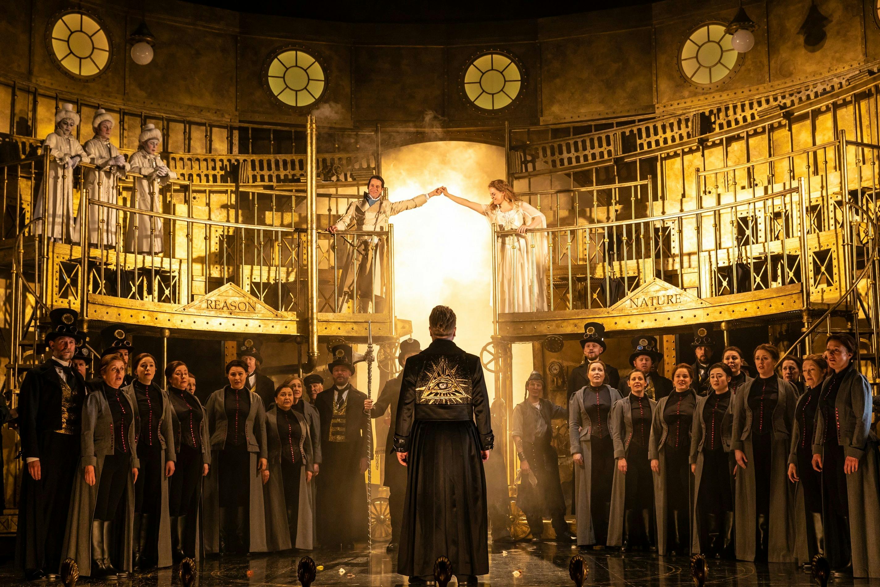 Ensemble in dark steampunk costumes surrounds two figures reaching across a glowing stage