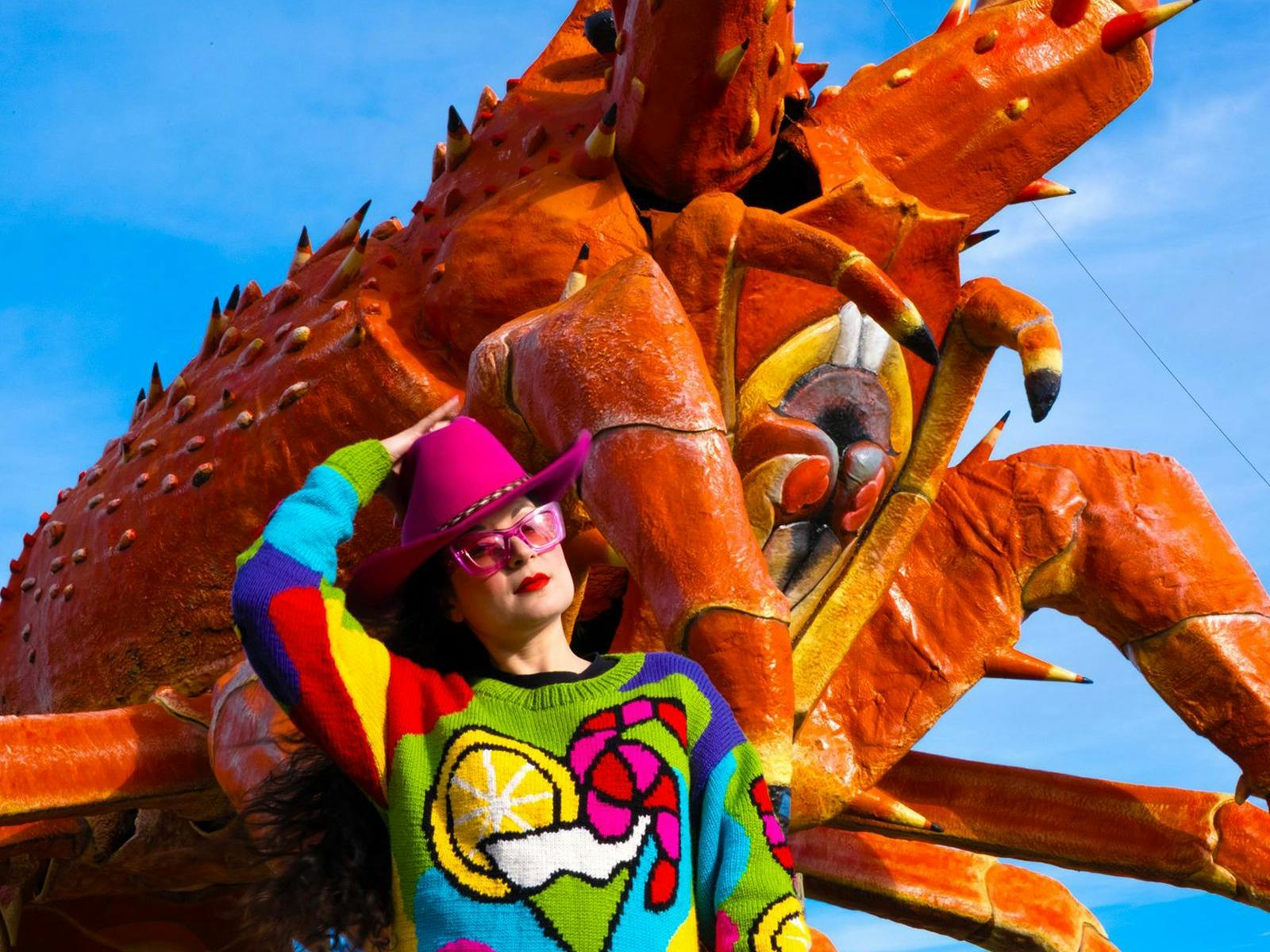 a woman in a colourful knitted jumper and pink cowboy hat in front of a giant orange crayfish