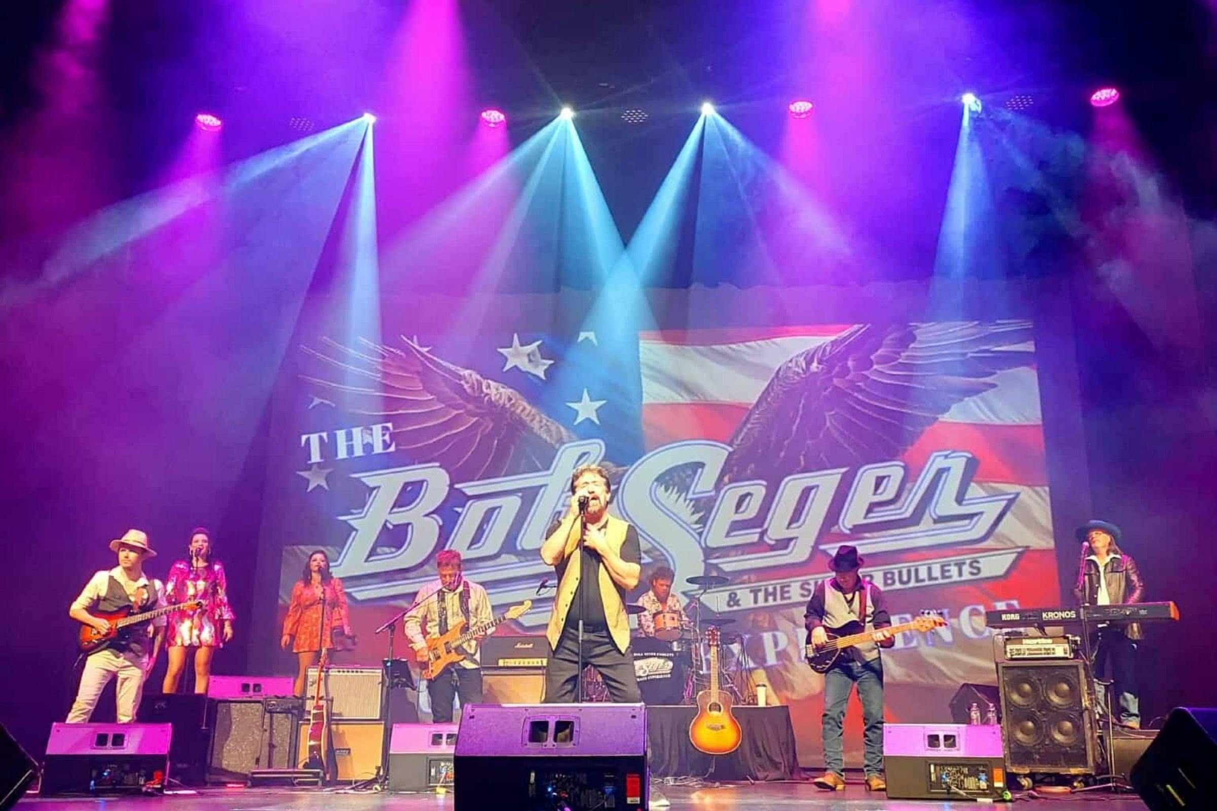 A rock band on stage, a large sign reads The Bob Seger & the Silver Bullets Ultimate Expereience