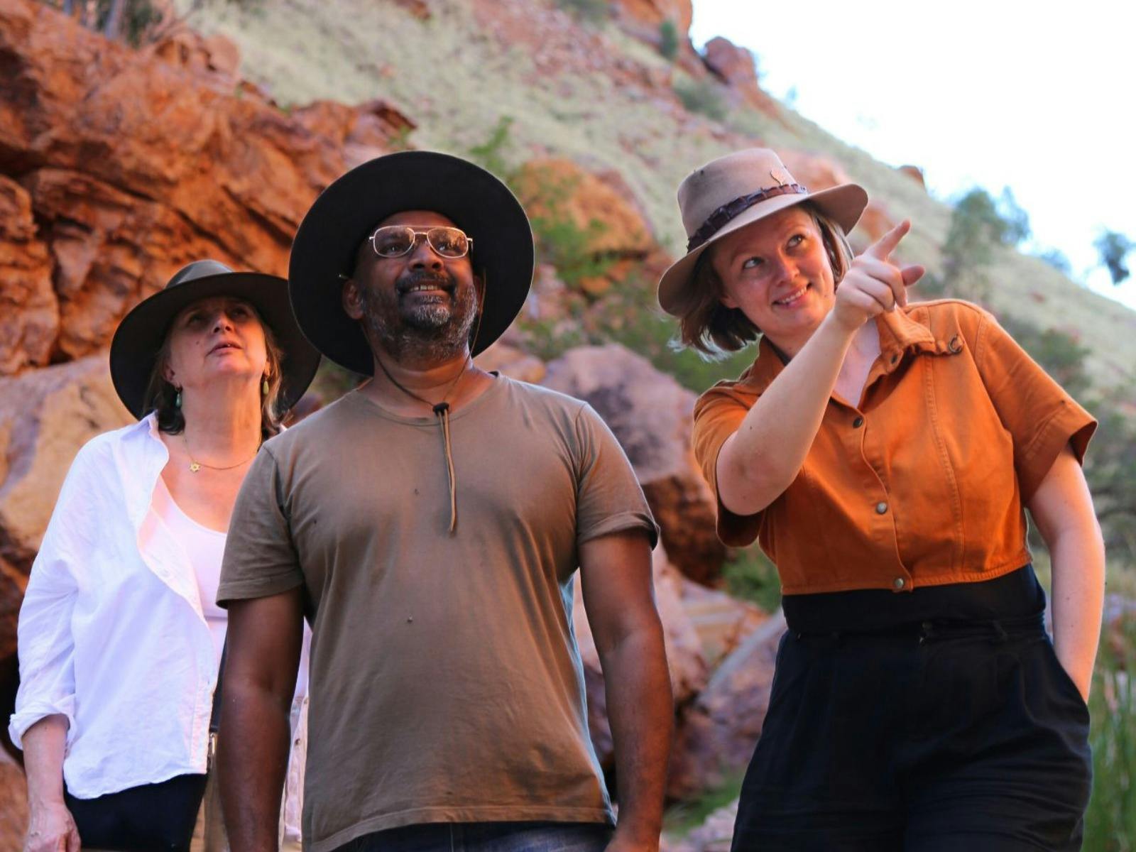 Three people stand in front of a rocky landscape, with one pointing at something in the distance.