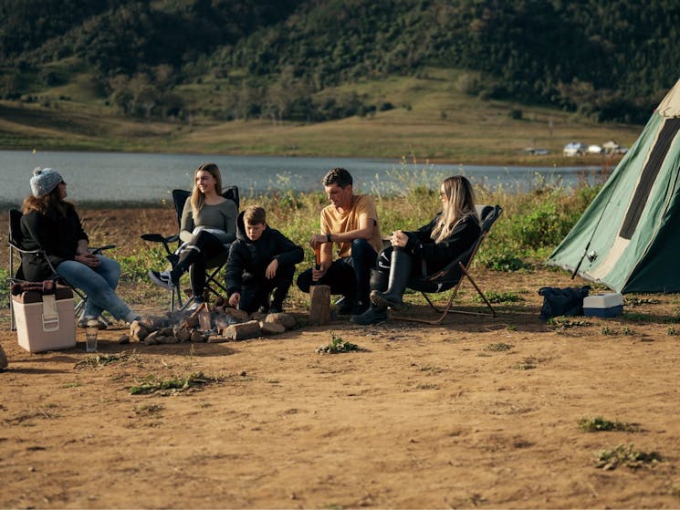 Family sits in camping chairs around a small campfire by the lake.