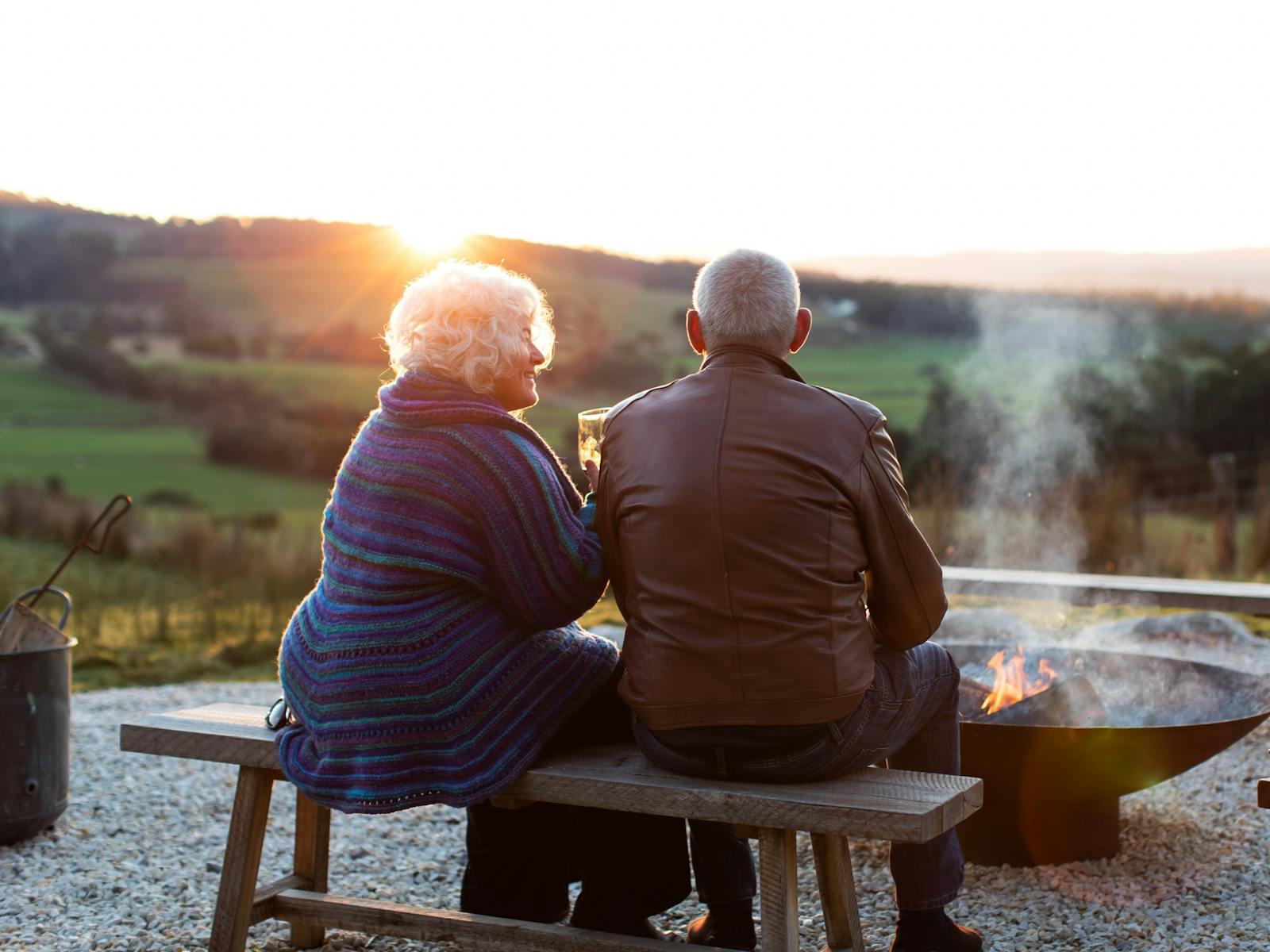 A couple sit close together on a wooden bench in front of a big fire pit, there is a valley view