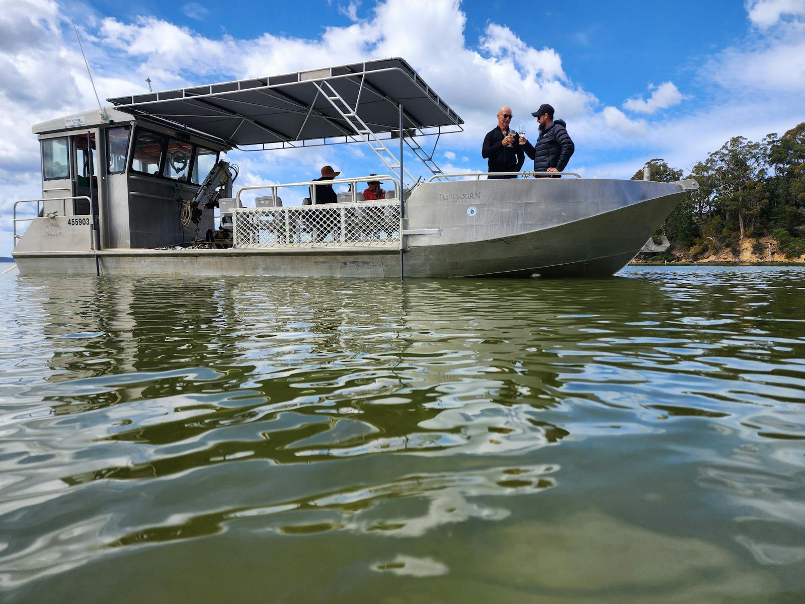 Boat glides on water with guests holding wine, shaded deck, leafy shoreline backdrop