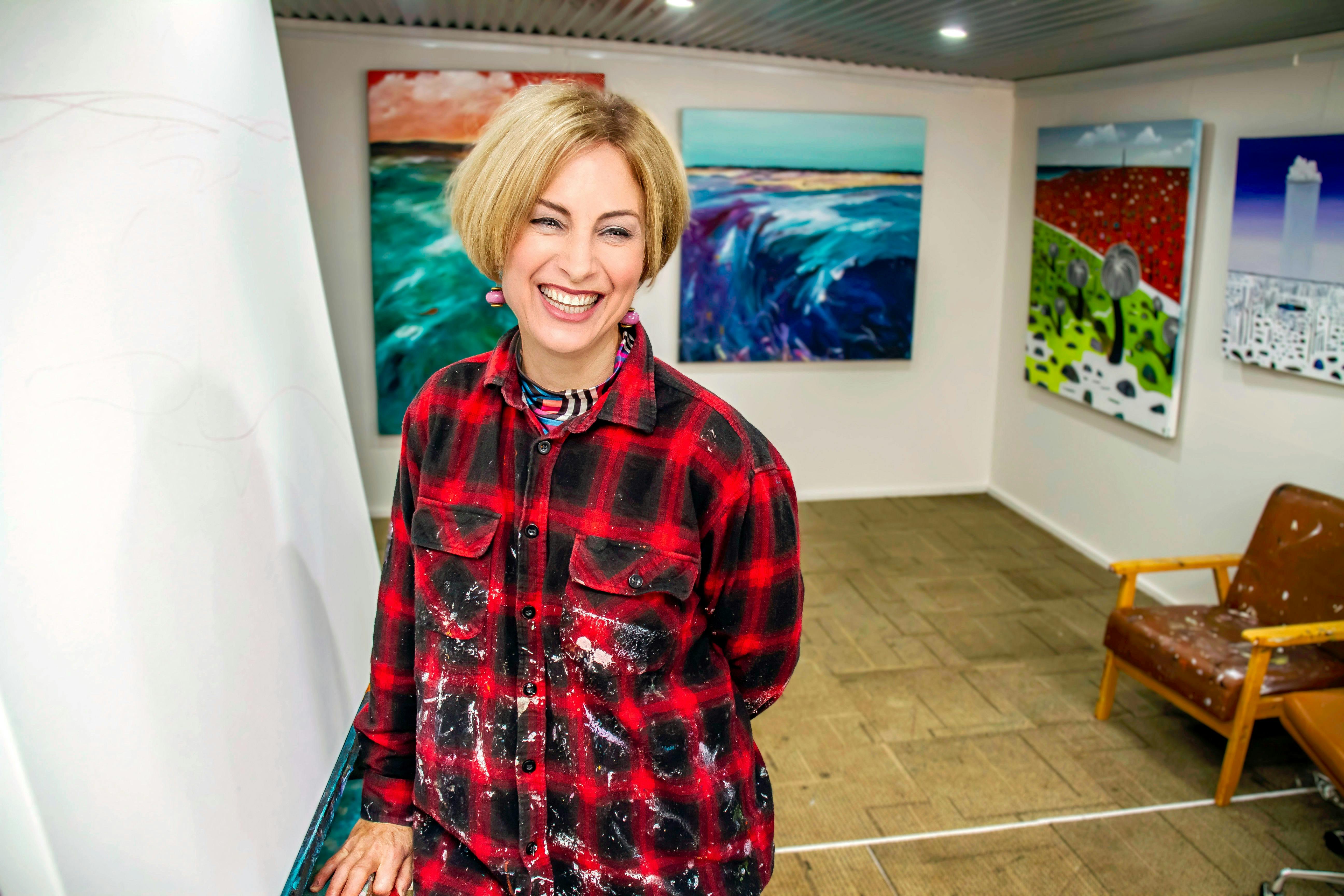 Smiling artist in a paint-splattered flannel shirt, standing in her studio surrounded by colourful o
