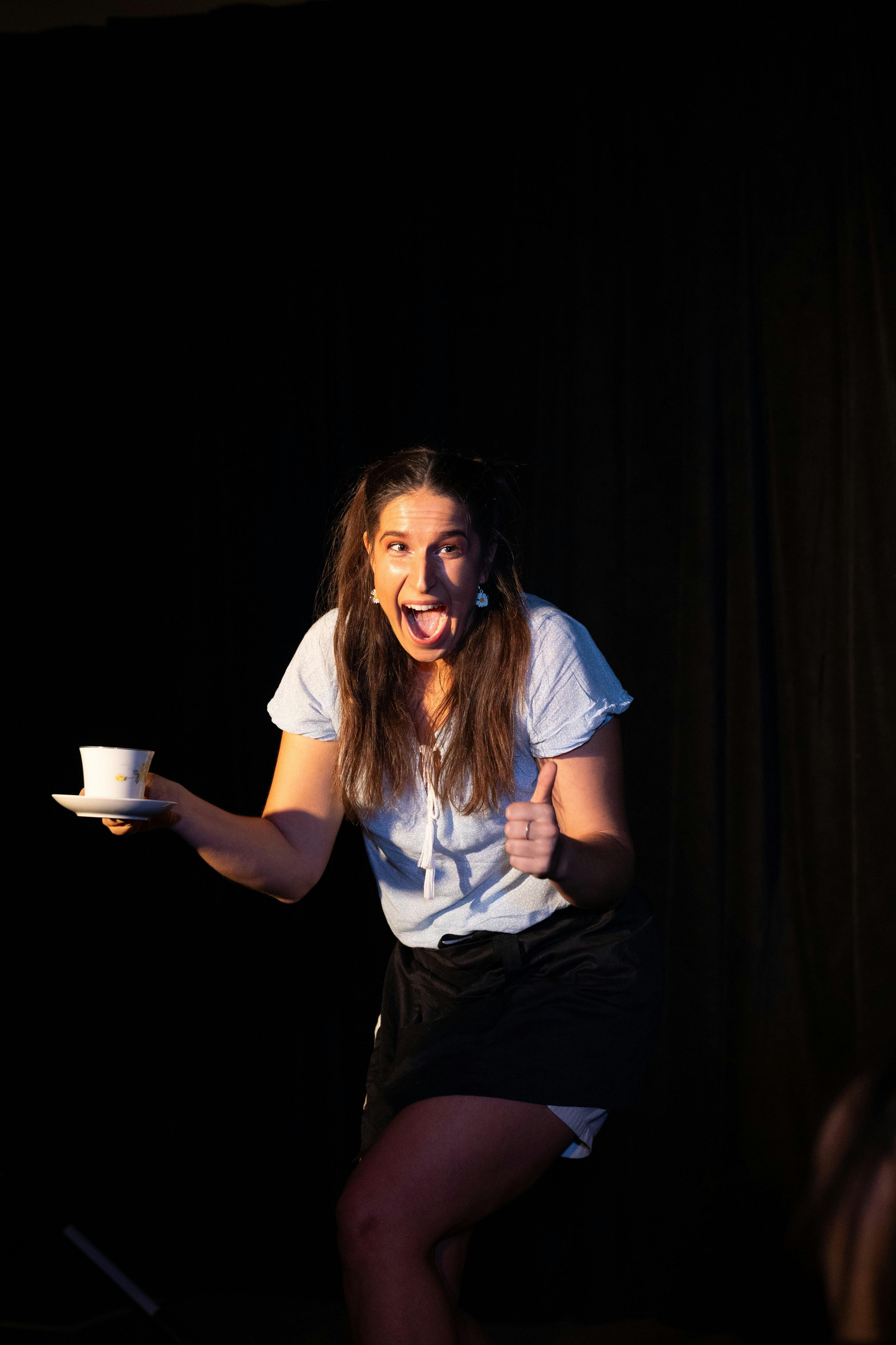 A waitress holding a coffee cup, giving a thumbs up. wearing a light blue shirt, black background.