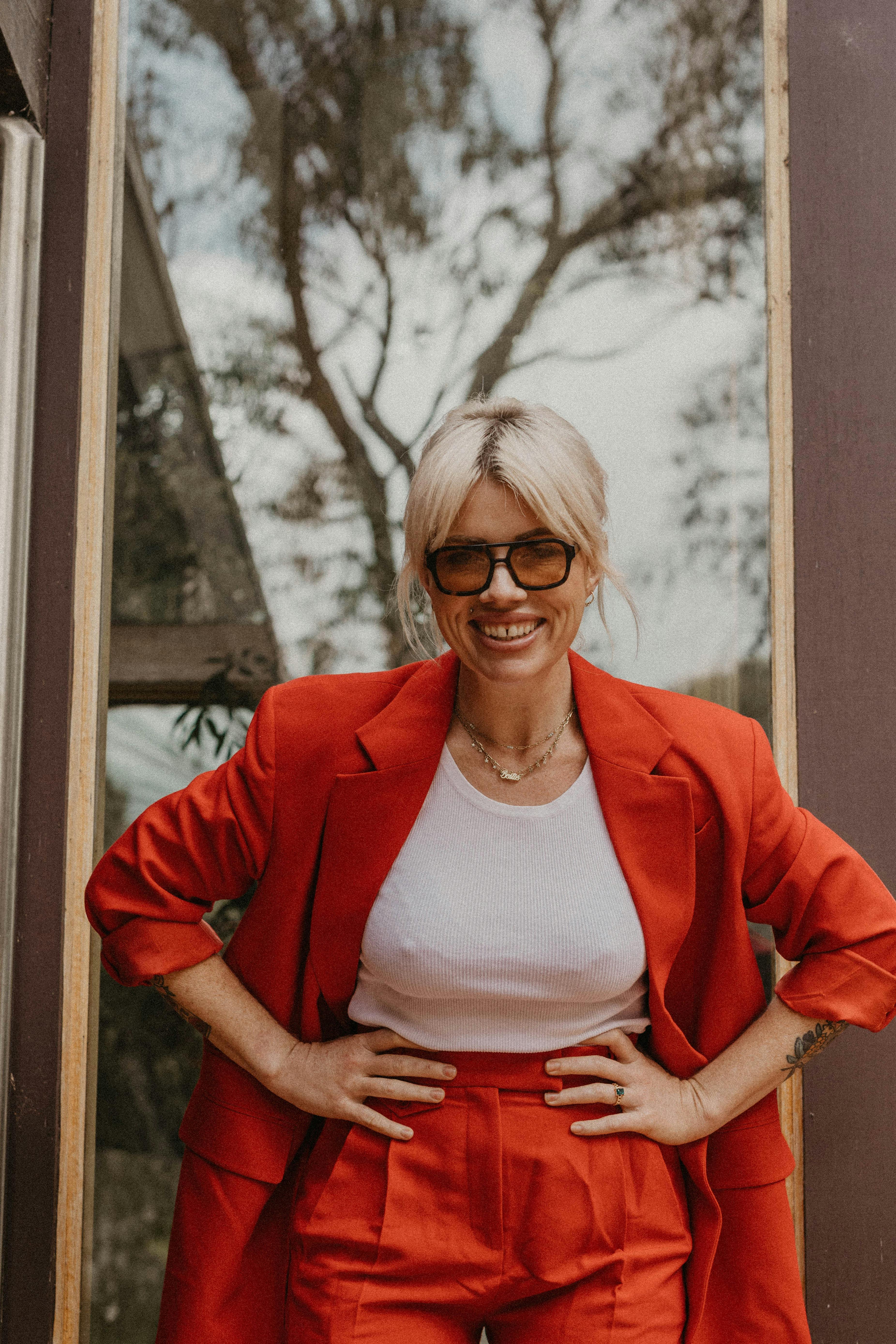 Clementine stands hands on hips in a red pantsuit, white shirt, trees reflected in the window behind