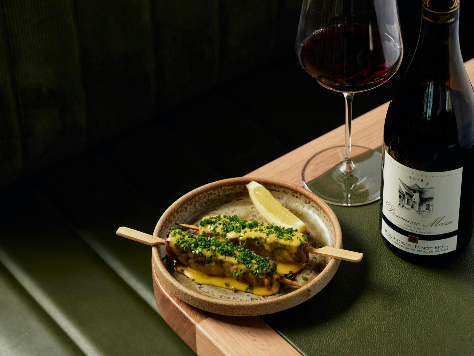 a plate filled with skewers sits on the corner of a green and pale wooden table next to some wine