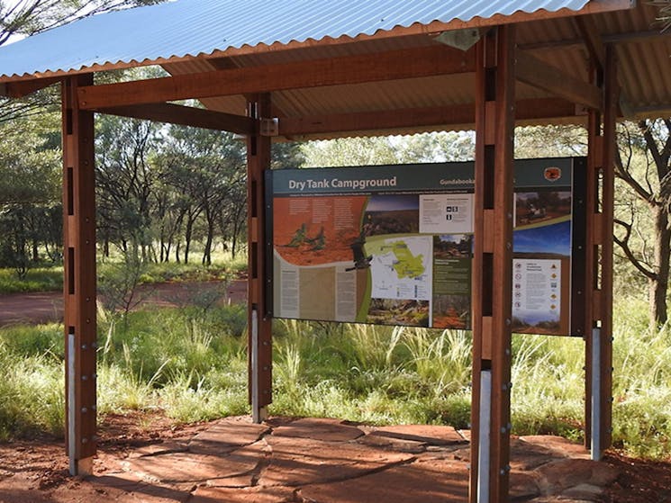 Photo of sign about Dry Tank campground under a shelter surrounded by trees, grass and red dirt at