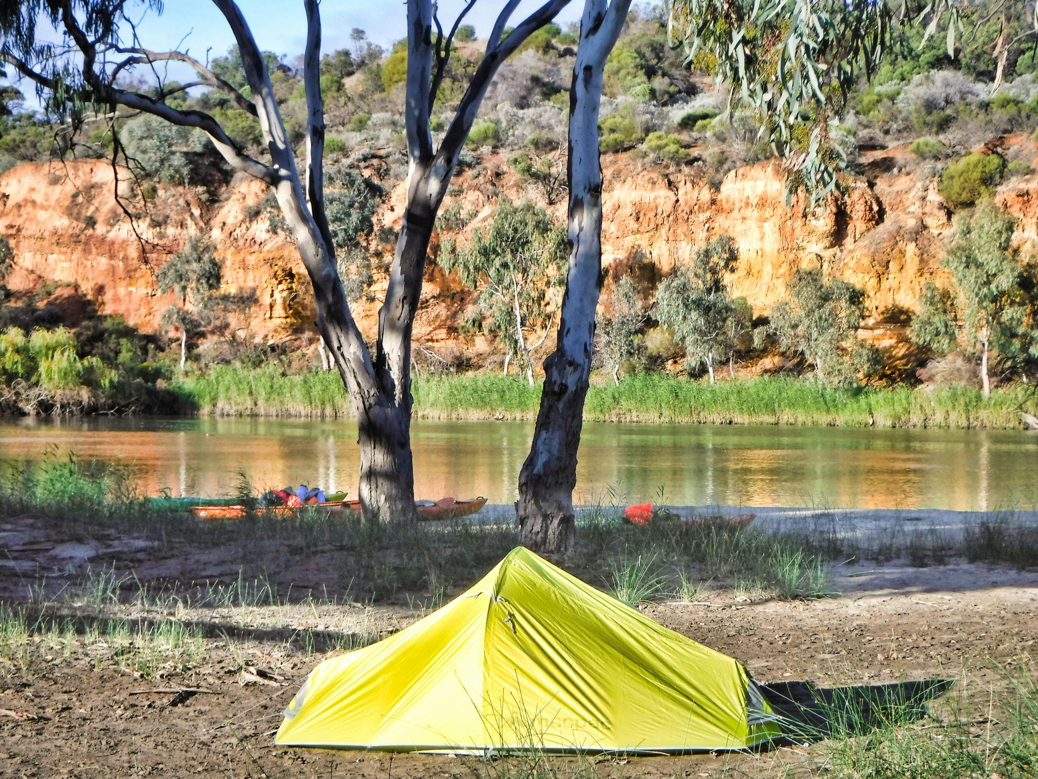 dome tent erected with background of kayaks, river and then lovely sandstone cliffs.