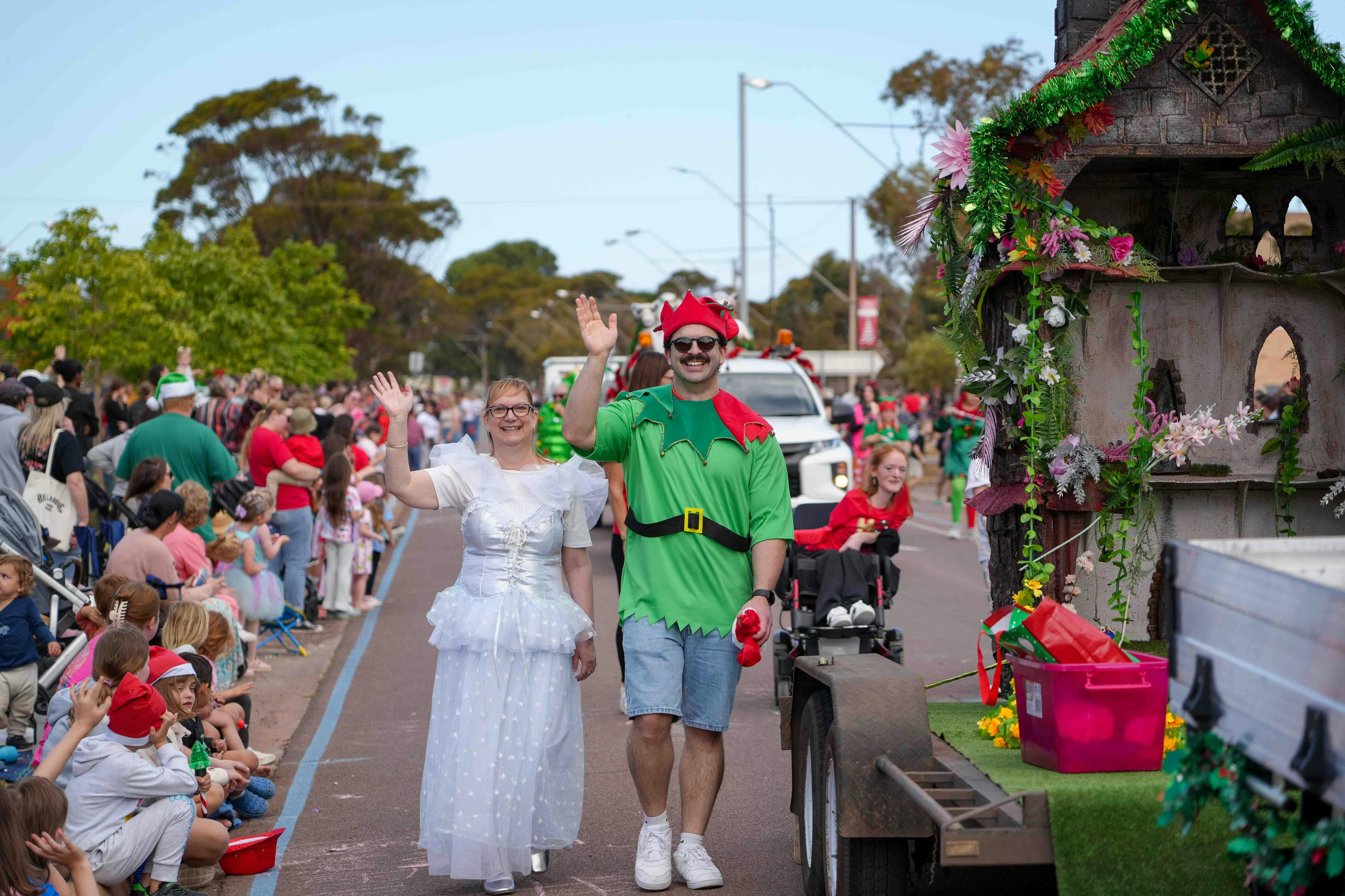A lady in a white fairy dress with a man dressed in an elfe outfit wavining to the crowd