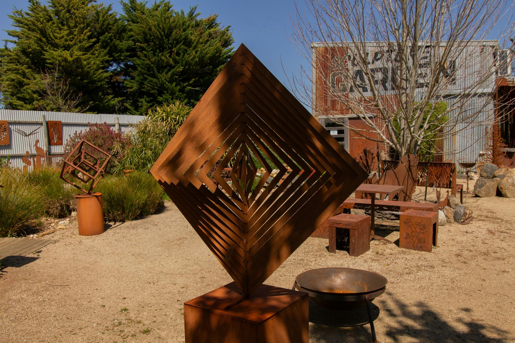 A metal square sculpture in the garden with bench seats and fire pit behind.