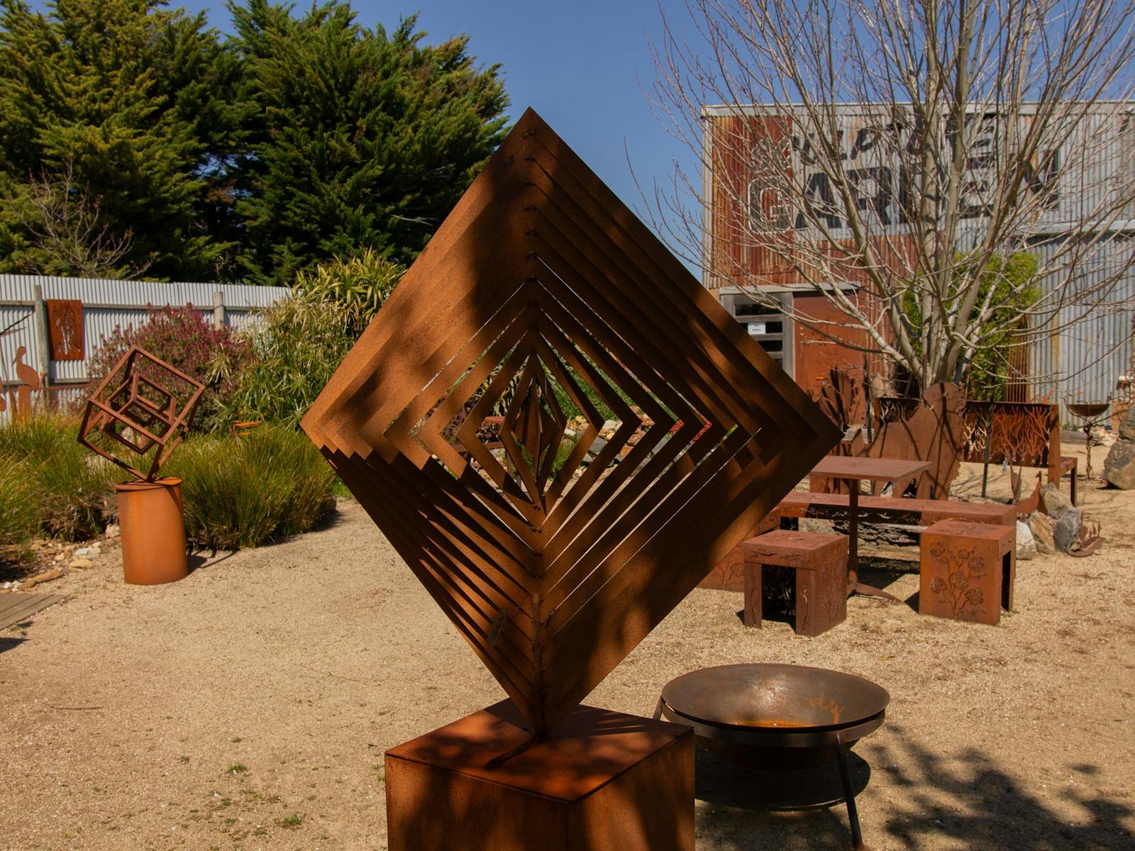A metal square sculpture in the garden with bench seats and fire pit behind.