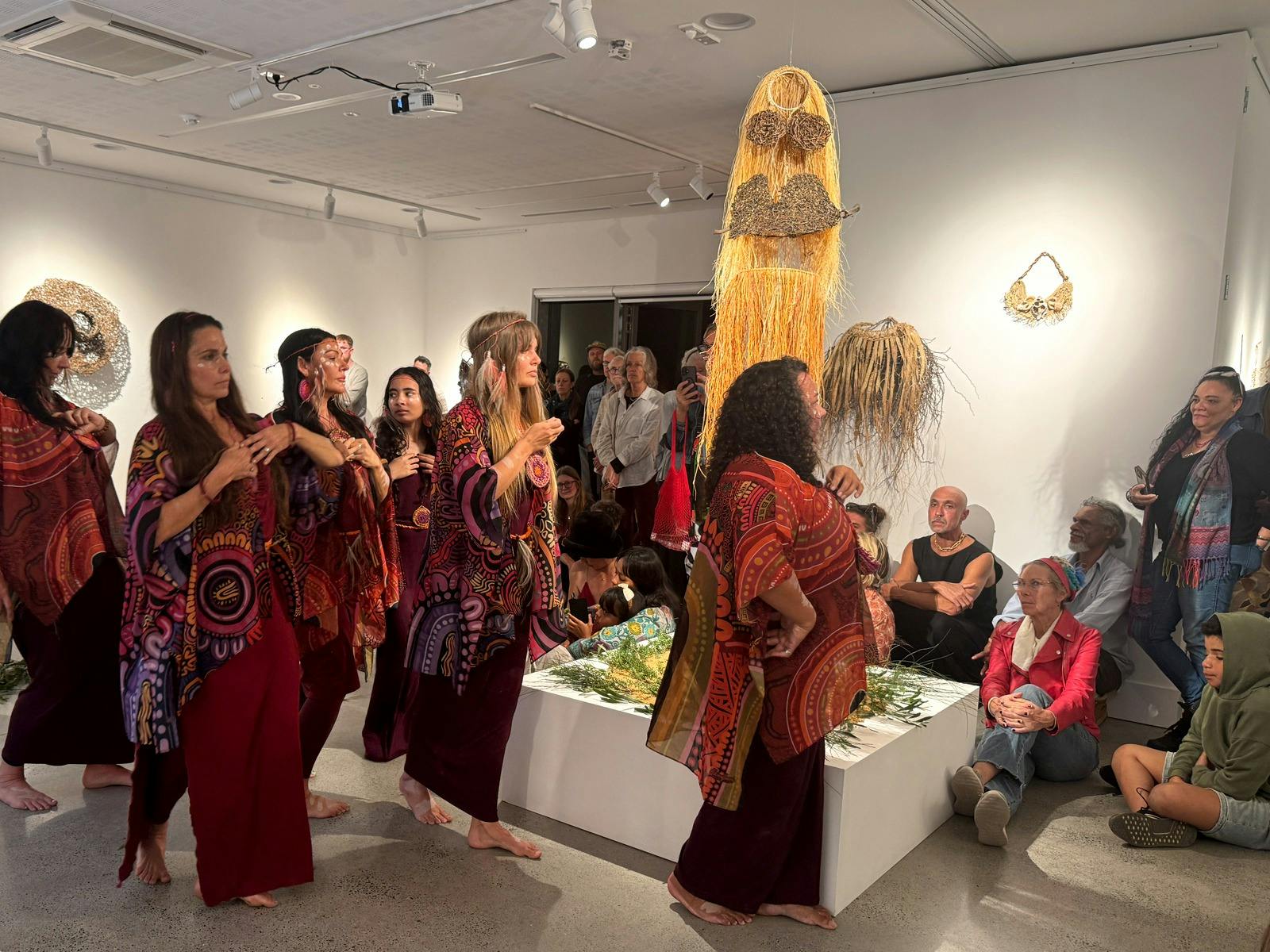 A group of Indigenous female dancers are moving in the gallery among some woven works and a crowd