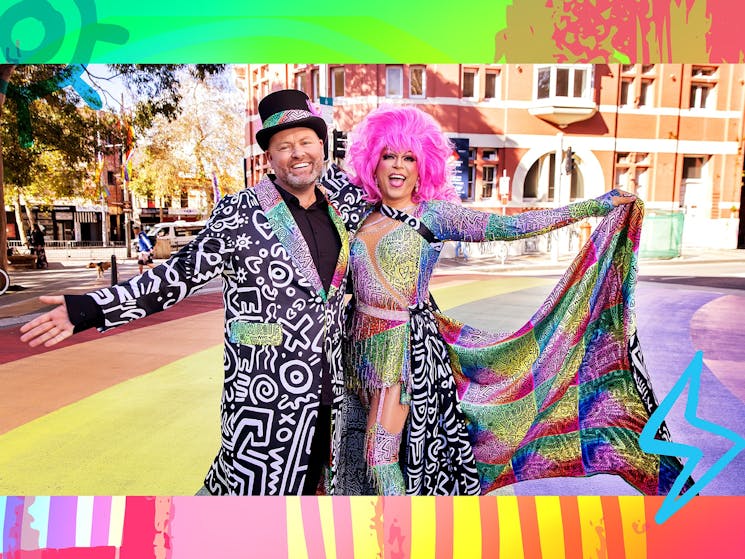 Two people in colourful rainbow outfits spreading their arms and smiling on a rainbow painted road