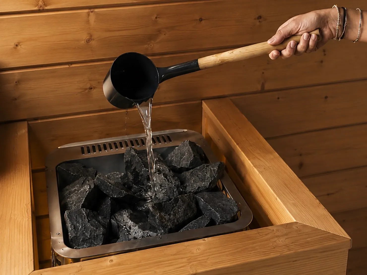 Hand pouring water from a ladle onto hot stones in a timber-lined Finnish sauna heater
