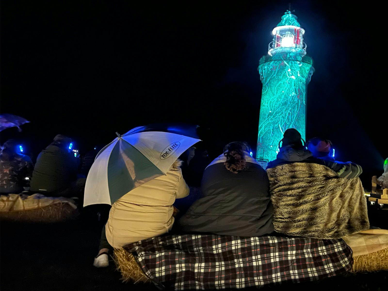 Peopl watching night time projections onto light house with an umbrella