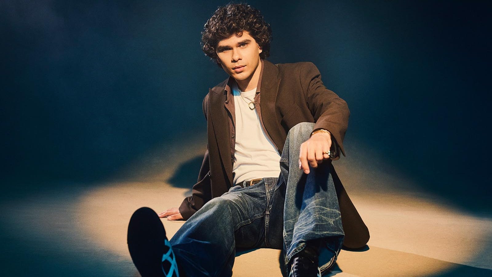 A young man with dark curly hair sitting on the floor in brown jacket and jeans A young man with dark curly hair sitting on the floor in brown jacket and jeans