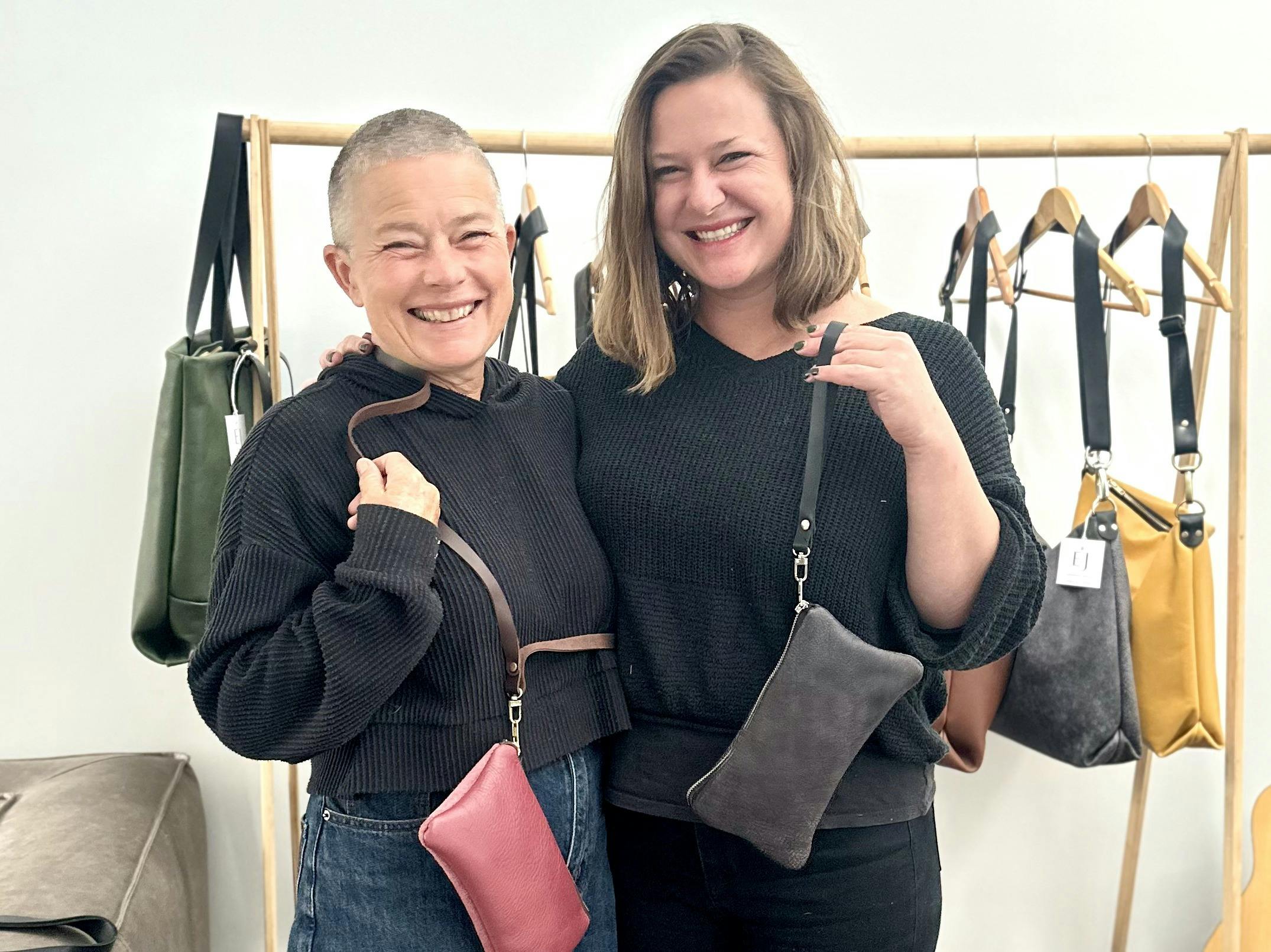 Two women smiling and holding their finished leather clutches with shoulder straps at a workshop, st