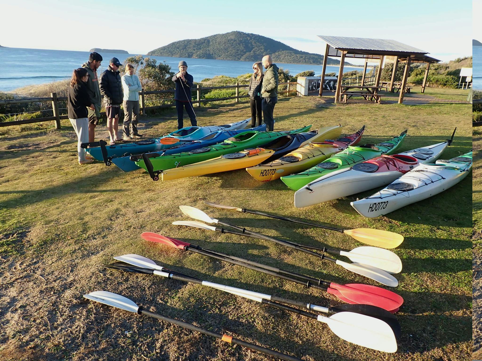 A number of Kayaks on the lawn with people having a lesson about them
