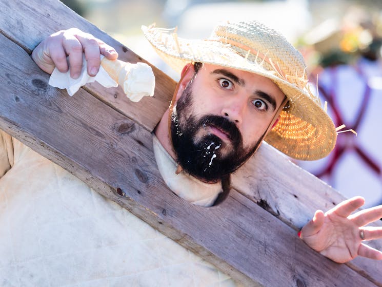 a bearded man wearing a straw hat is contained in stocks, holding an icecream in his right hand