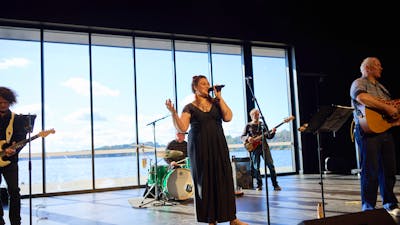 A woman sings passionately on a stage in front of a windowed-wall looking onto Lake Ginninderra.