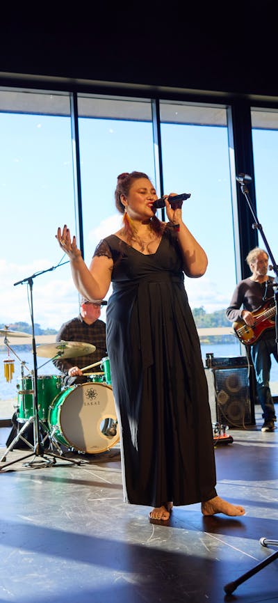 A woman sings passionately on a stage in front of a windowed-wall looking onto Lake Ginninderra.