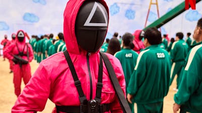 A still shot from movie Squid Game. Men in pink and green suits.