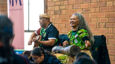 2 performers play music for an audience, one playing a guitar and one playing percussion instrument