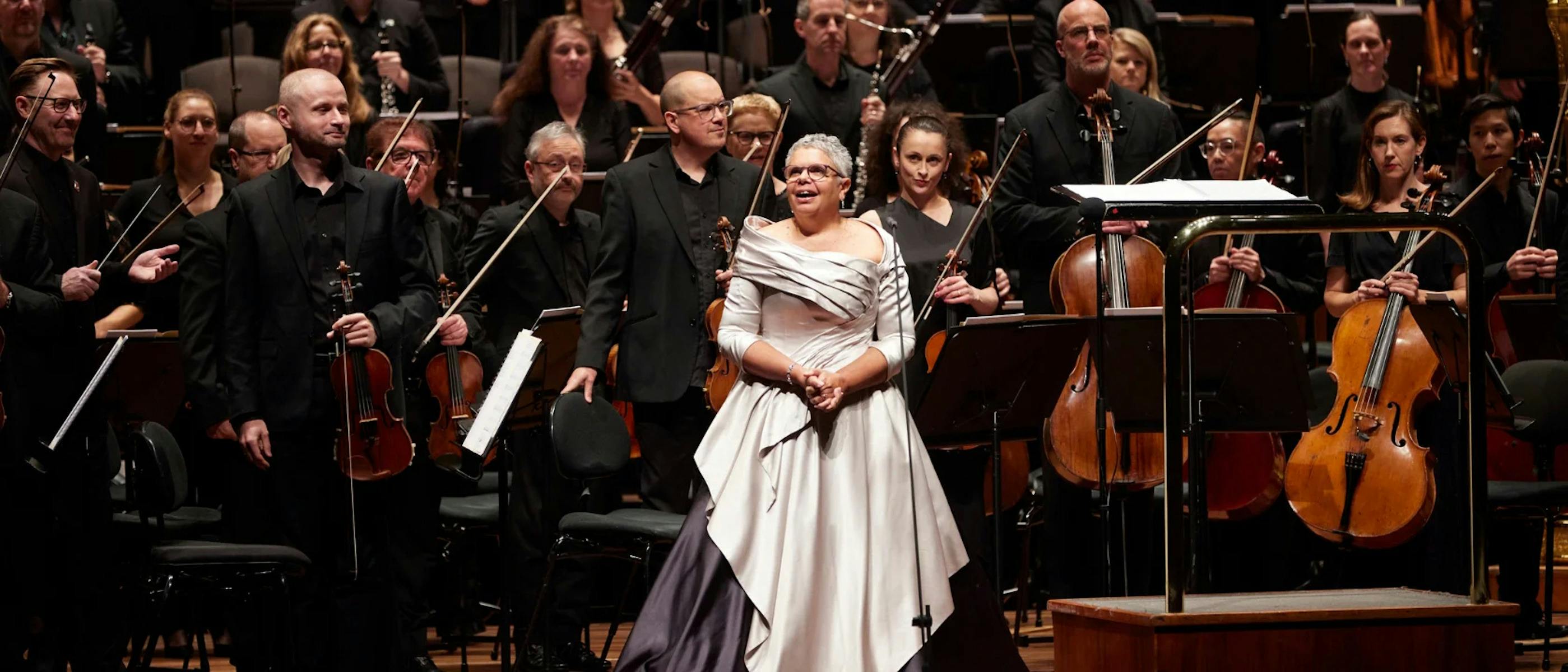 lady dressed in siver smiling on stage in front of an orchaestra