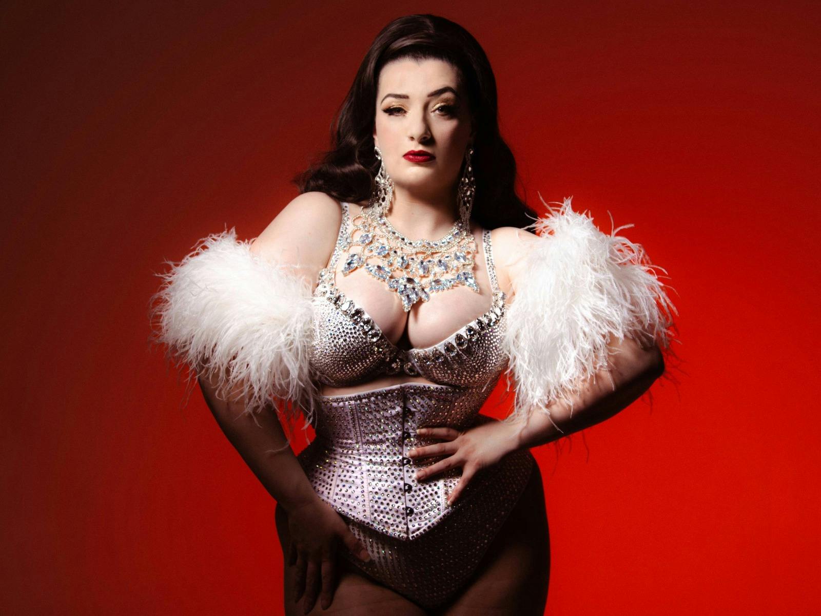 Connie is wearing a sparkly corset and feather arm cuffs. She is posing with hand on her hips
