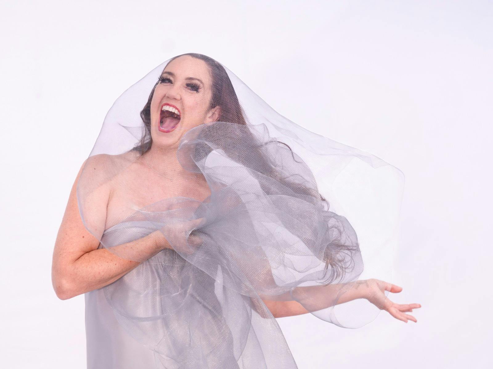 A woman stands in a grey veiled dress screaming at the camera
